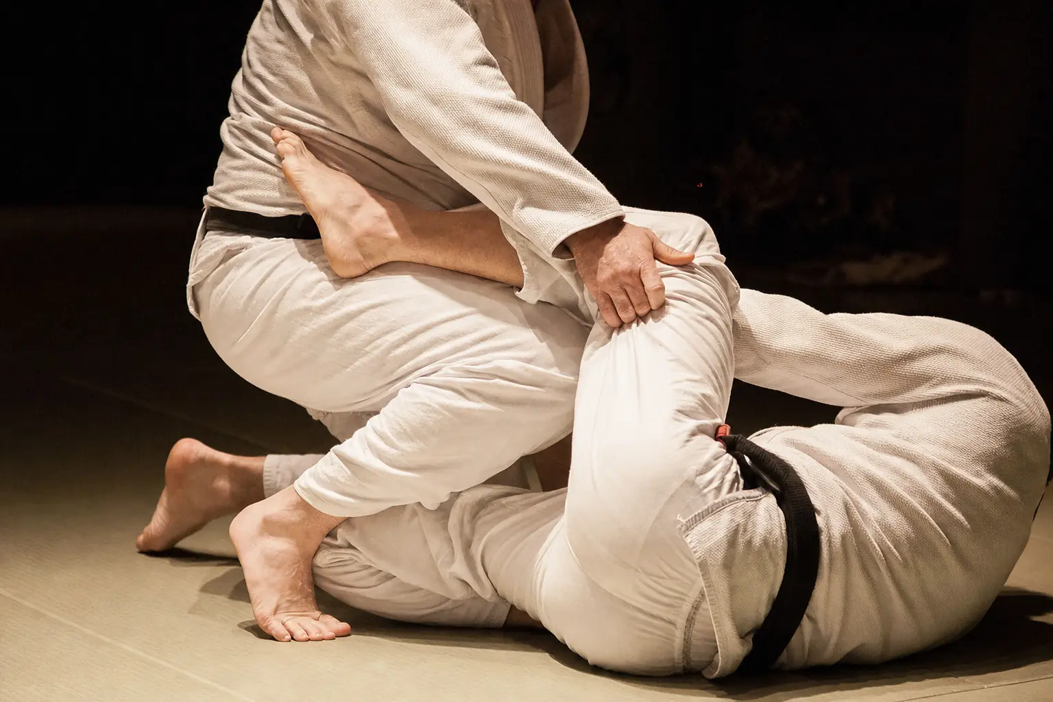 Deux pratiquants en kimono, ceintures noires, au corps à corps sur un tatami.